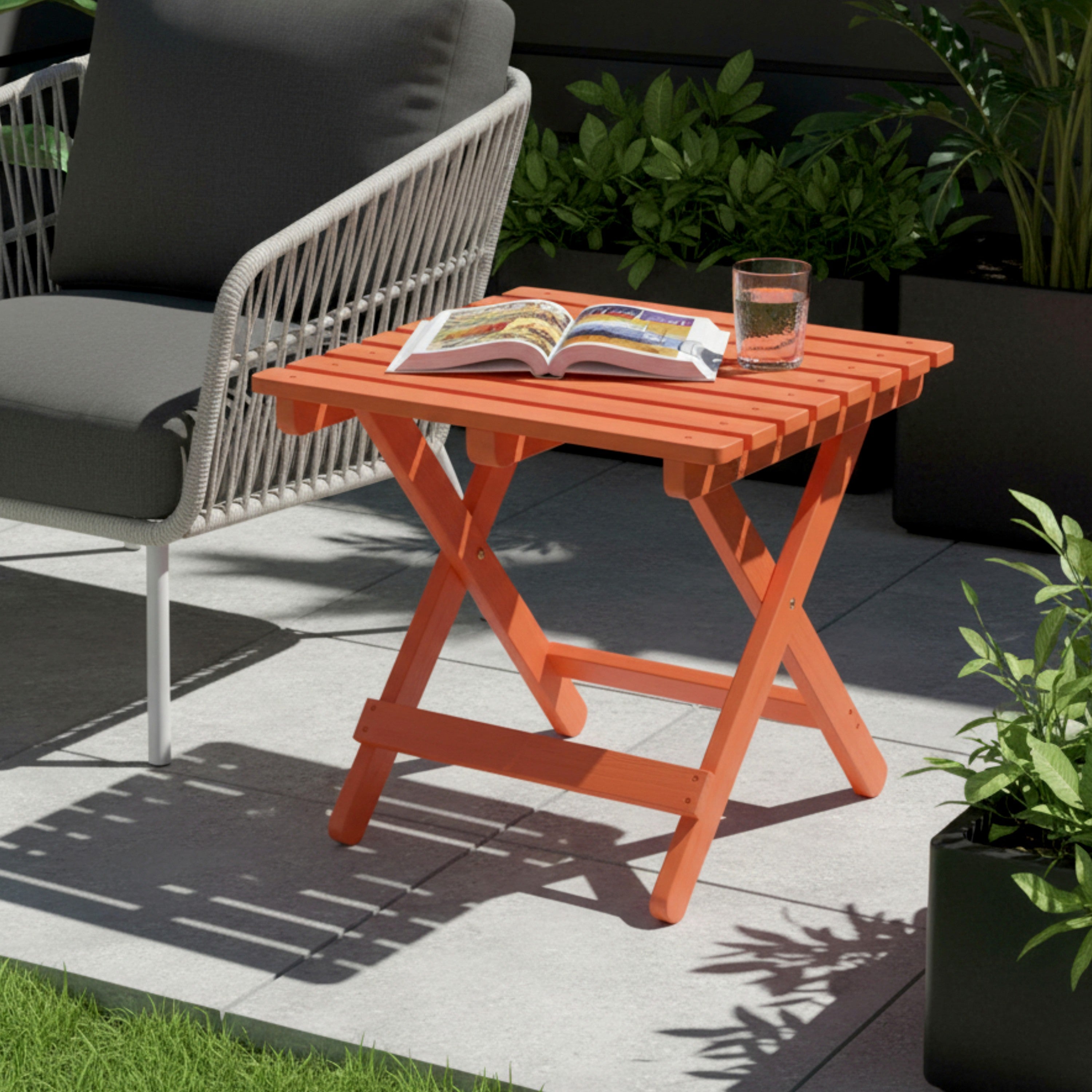 An orange folding side table in an outdoor patio setting, styled with an open magazine and a glass of water on top, next to a modern white woven armchair. 