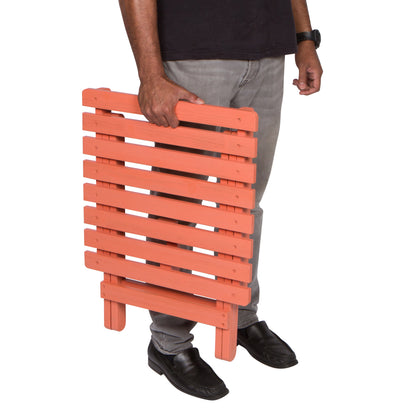 A person holding a portable folding wooden side table with orange-painted slats, shown from the waist down against a plain white background. 