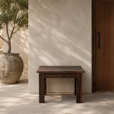 A dark brown wooden bench with a slatted top and block legs, positioned against a textured beige stucco wall with dappled sunlight and shadows. A large rustic planter with a tree is on the left, and a dark wood door is on the right. 