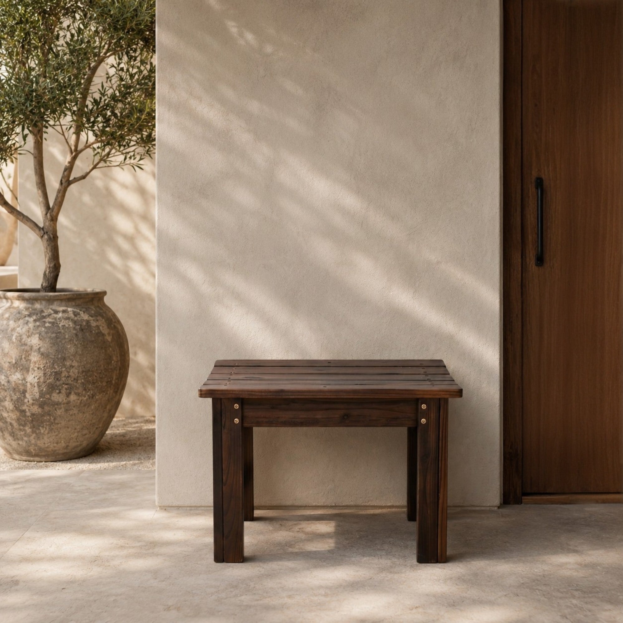 A dark brown wooden bench with a slatted top and block legs, positioned against a textured beige stucco wall with dappled sunlight and shadows. A large rustic planter with a tree is on the left, and a dark wood door is on the right. 