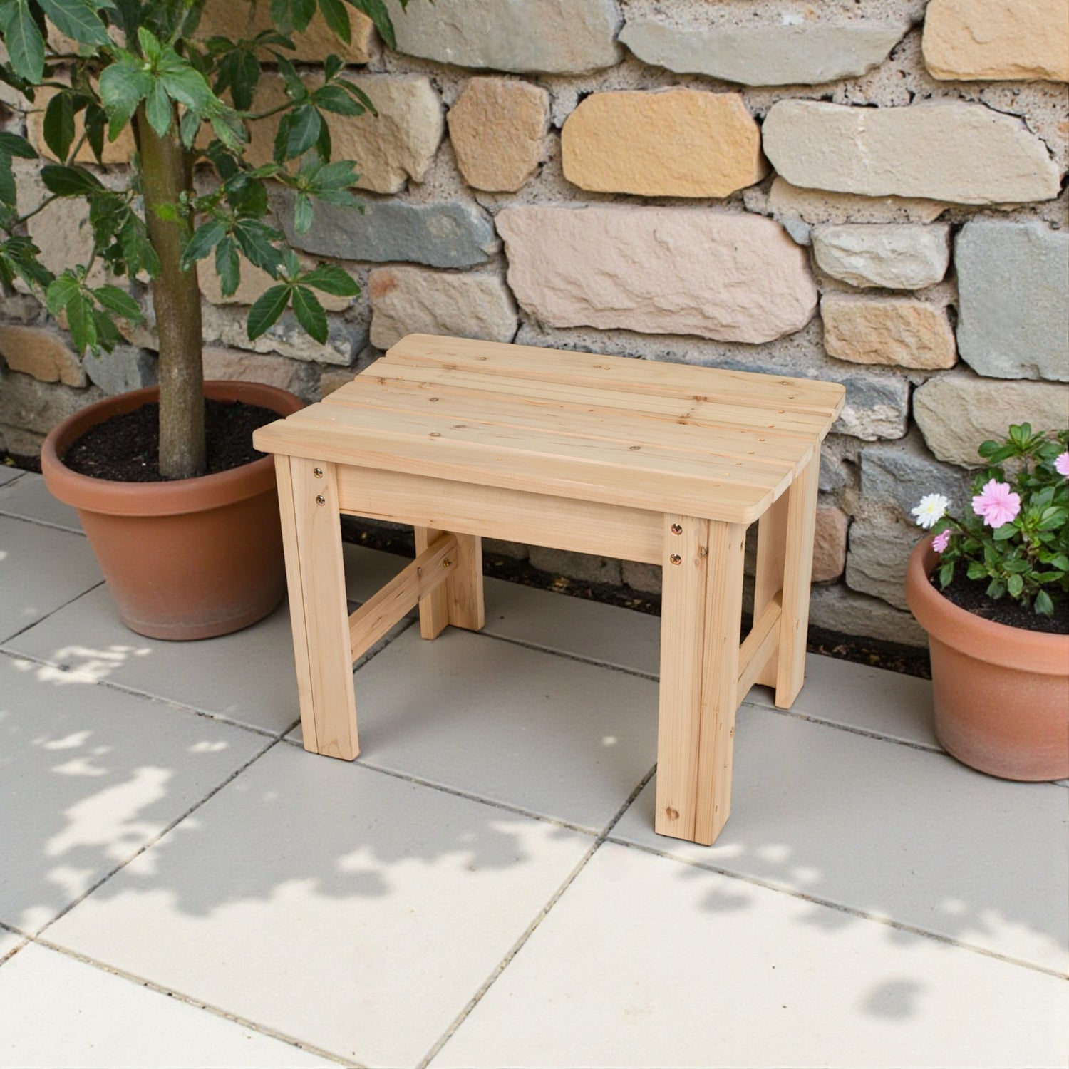 A small rectangular wooden bench or stool made of light natural wood, placed on a tiled patio in front of a stone wall with potted plants on both sides. 