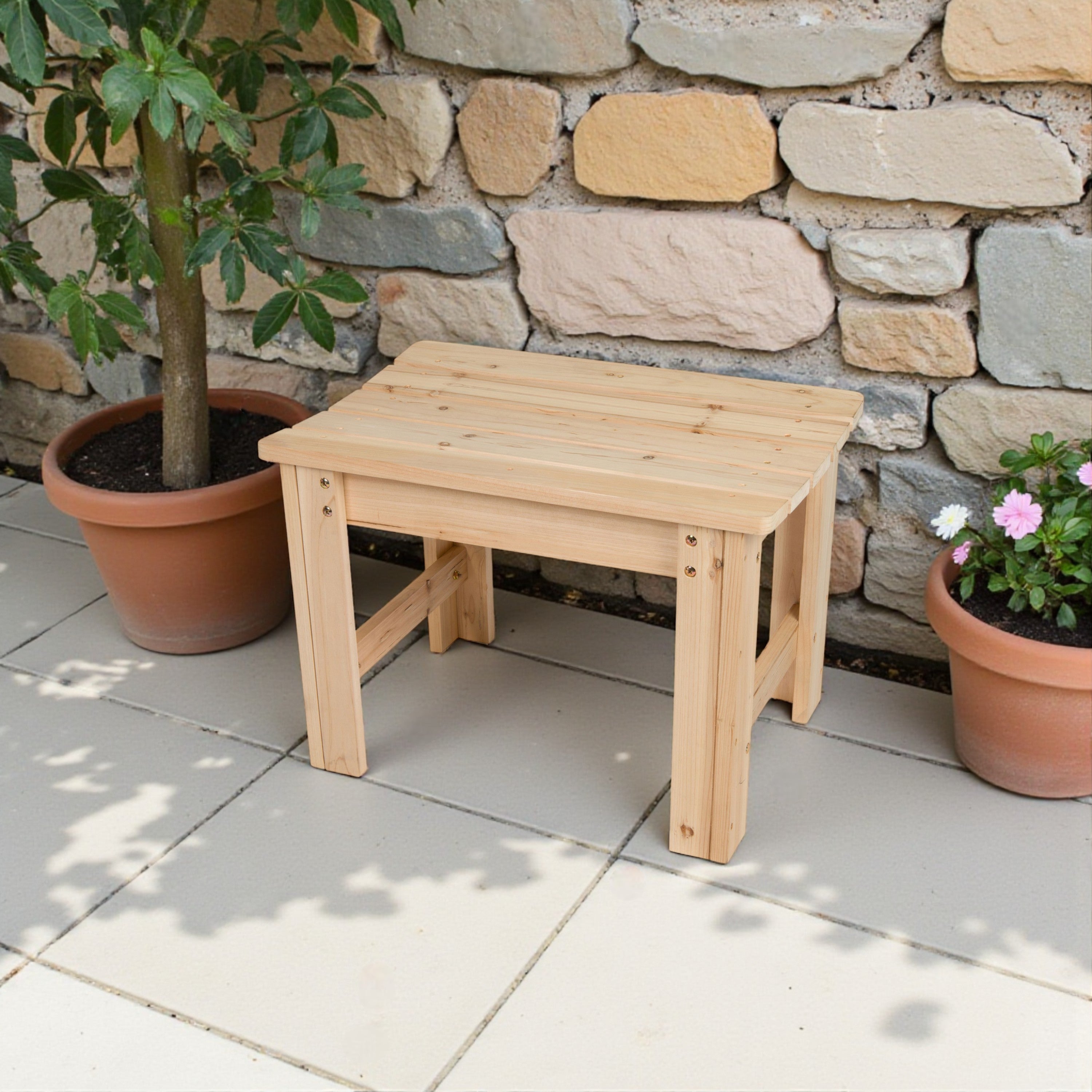 A small rectangular wooden bench or stool made of light natural wood, placed on a tiled patio in front of a stone wall with potted plants on both sides. 