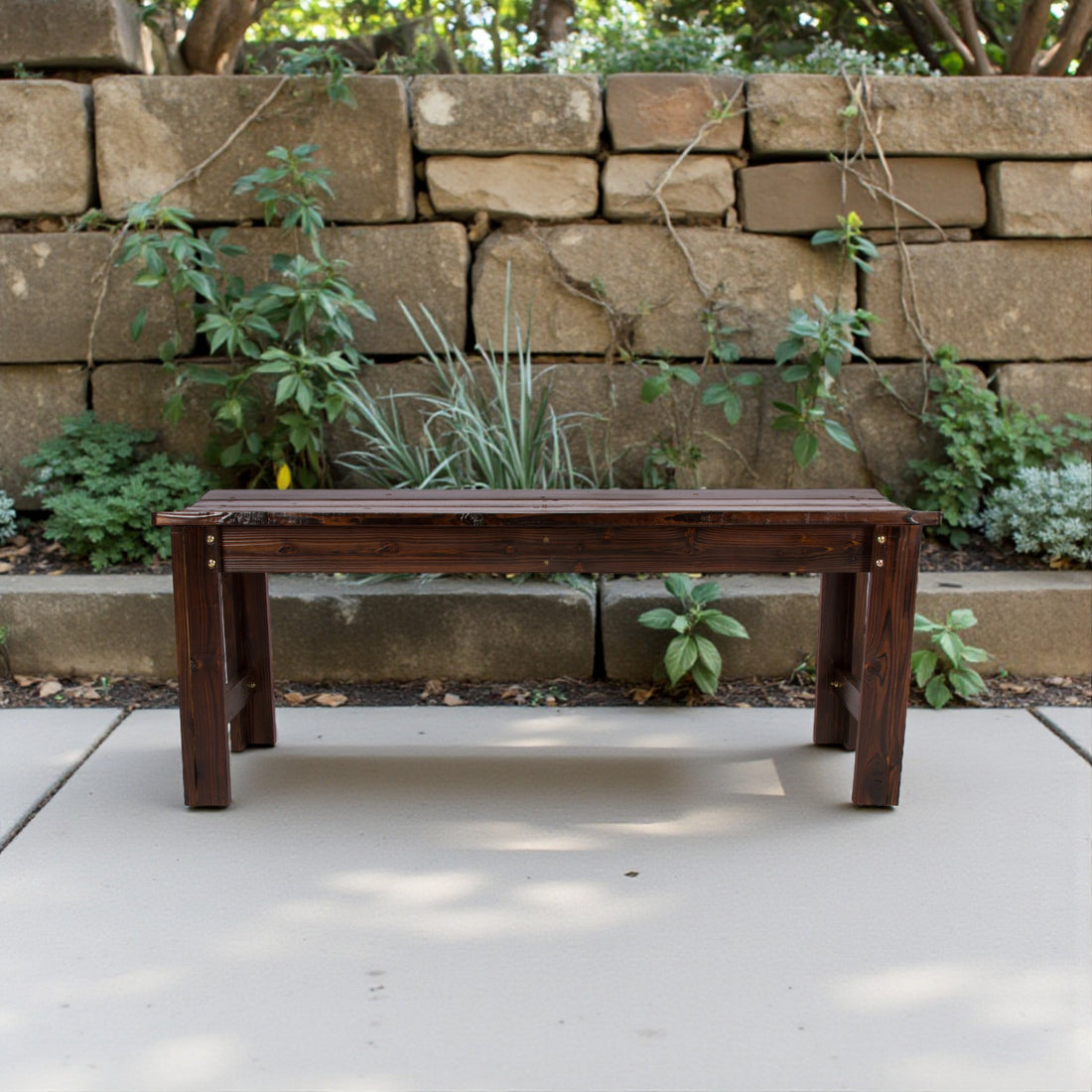 A dark brown wooden bench with a rustic, weathered finish sits on a light concrete patio in front of a stacked stone wall with green plants. 