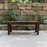 A dark brown wooden bench with a rustic, weathered finish sits on a light concrete patio in front of a stacked stone wall with green plants. 