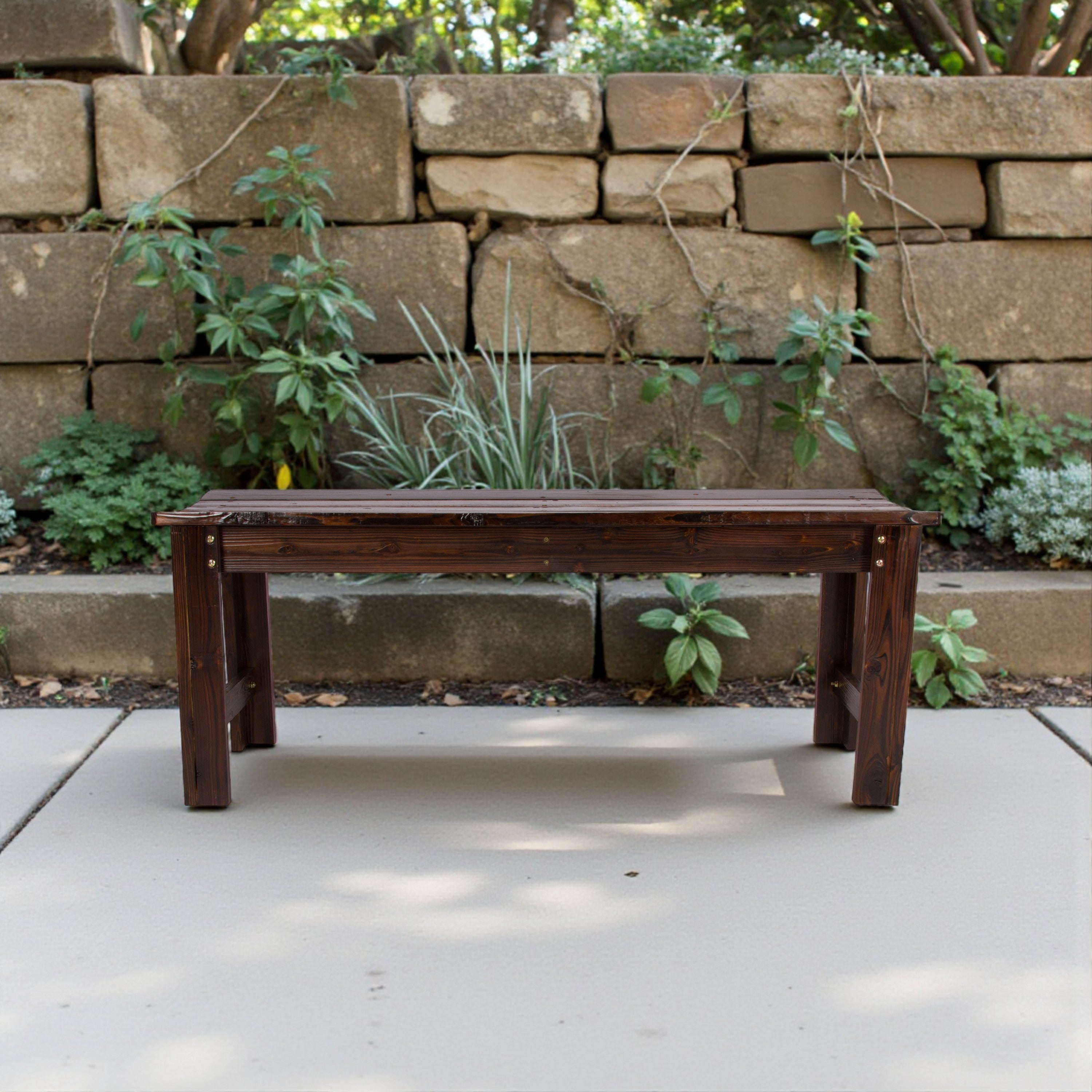 A dark brown wooden bench with a rustic, weathered finish sits on a light concrete patio in front of a stacked stone wall with green plants. 