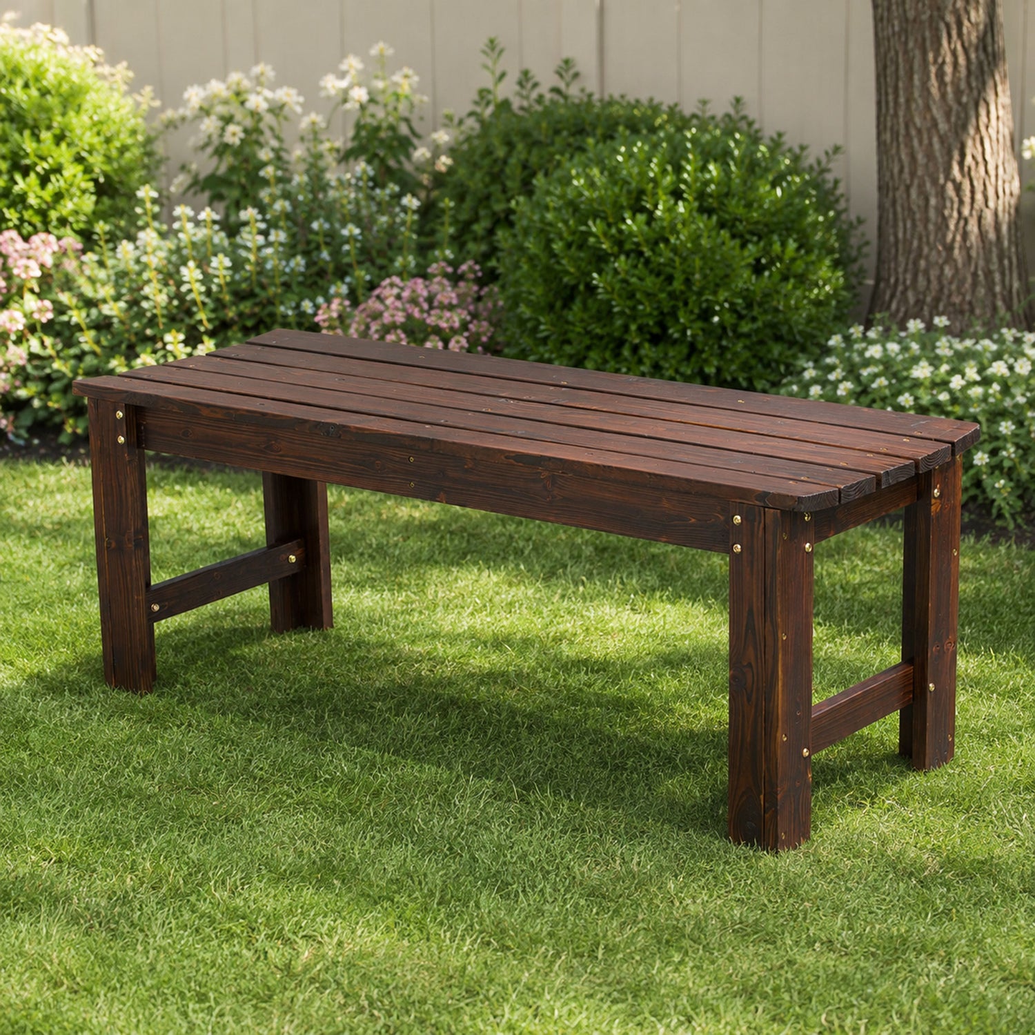 A dark brown wooden garden bench with a slatted top and sturdy legs, positioned on a green lawn in front of a garden with bushes and a fence. 