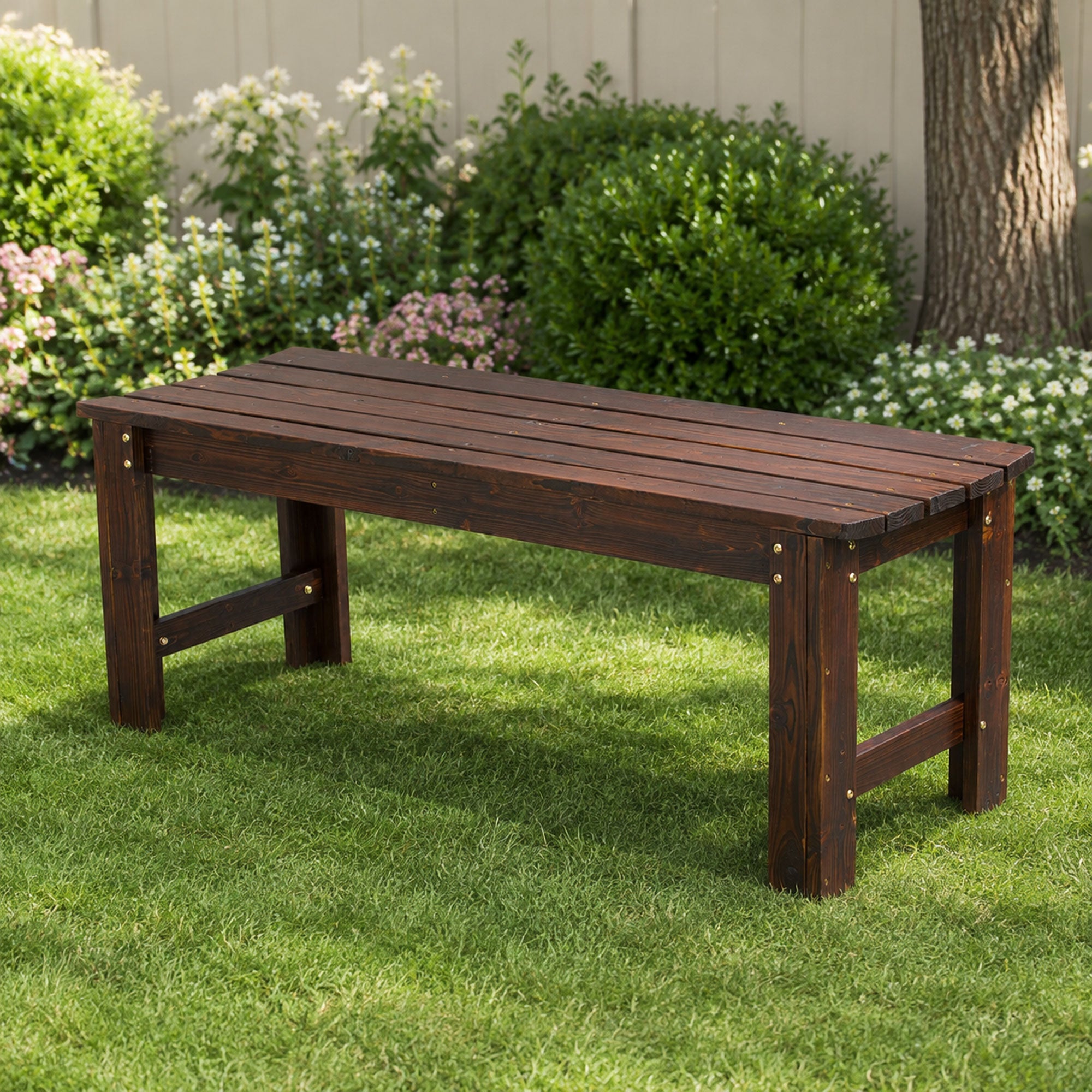 A dark brown wooden garden bench with a slatted top and sturdy legs, positioned on a green lawn in front of a garden with bushes and a fence. 