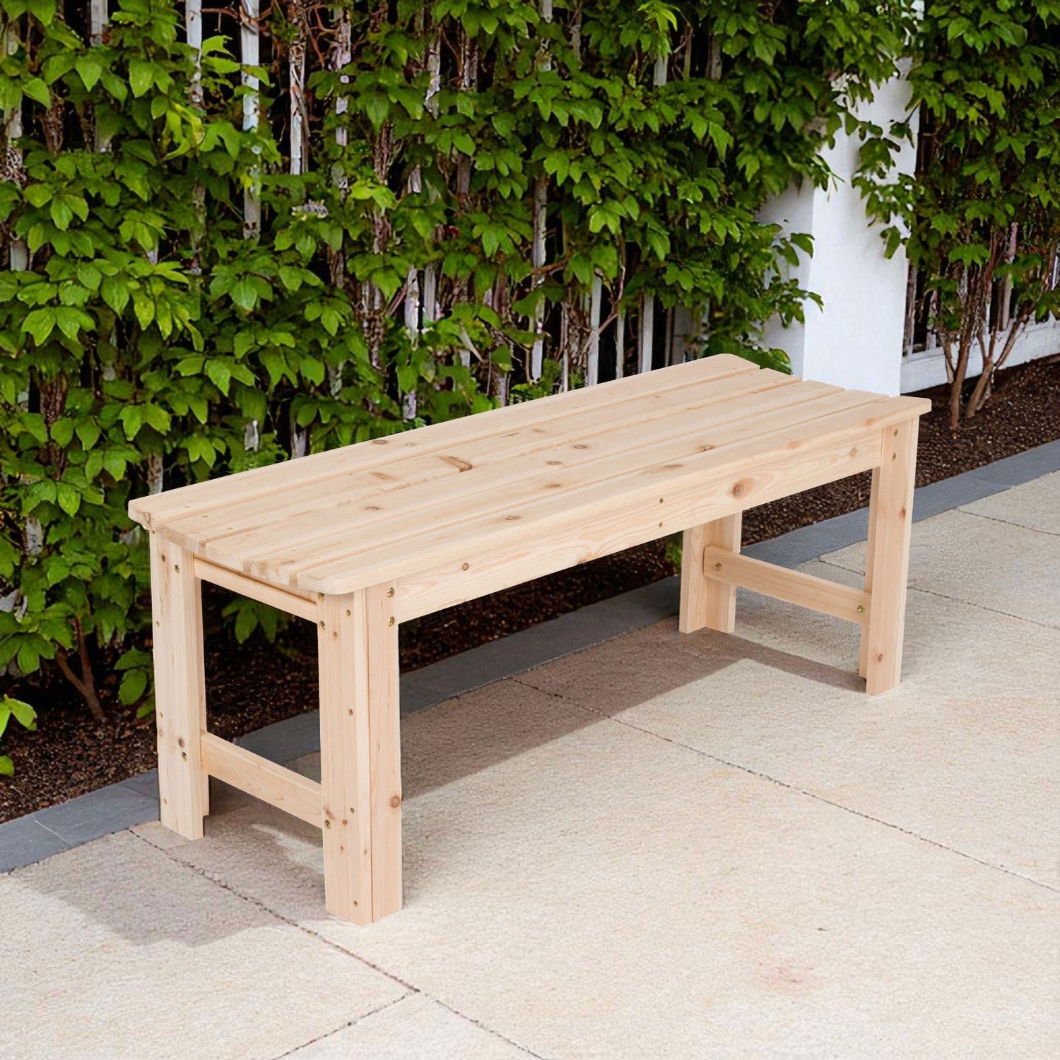 A light natural wood outdoor bench with a slatted seat and sturdy legs, positioned on a paved patio in front of a green leafy hedge. 