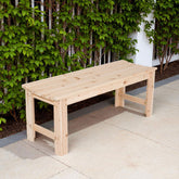 A light natural wood outdoor bench with a slatted seat and sturdy legs, positioned on a paved patio in front of a green leafy hedge. 