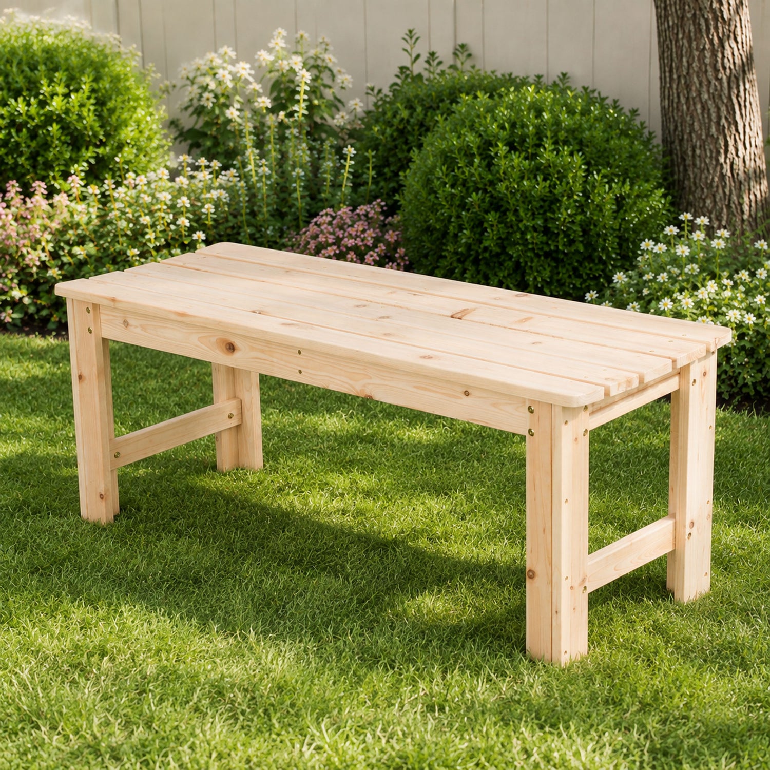 A light natural wood garden bench with a slatted top and sturdy legs, positioned on a green grass lawn with a garden fence, bushes, and a tree trunk in the background. 