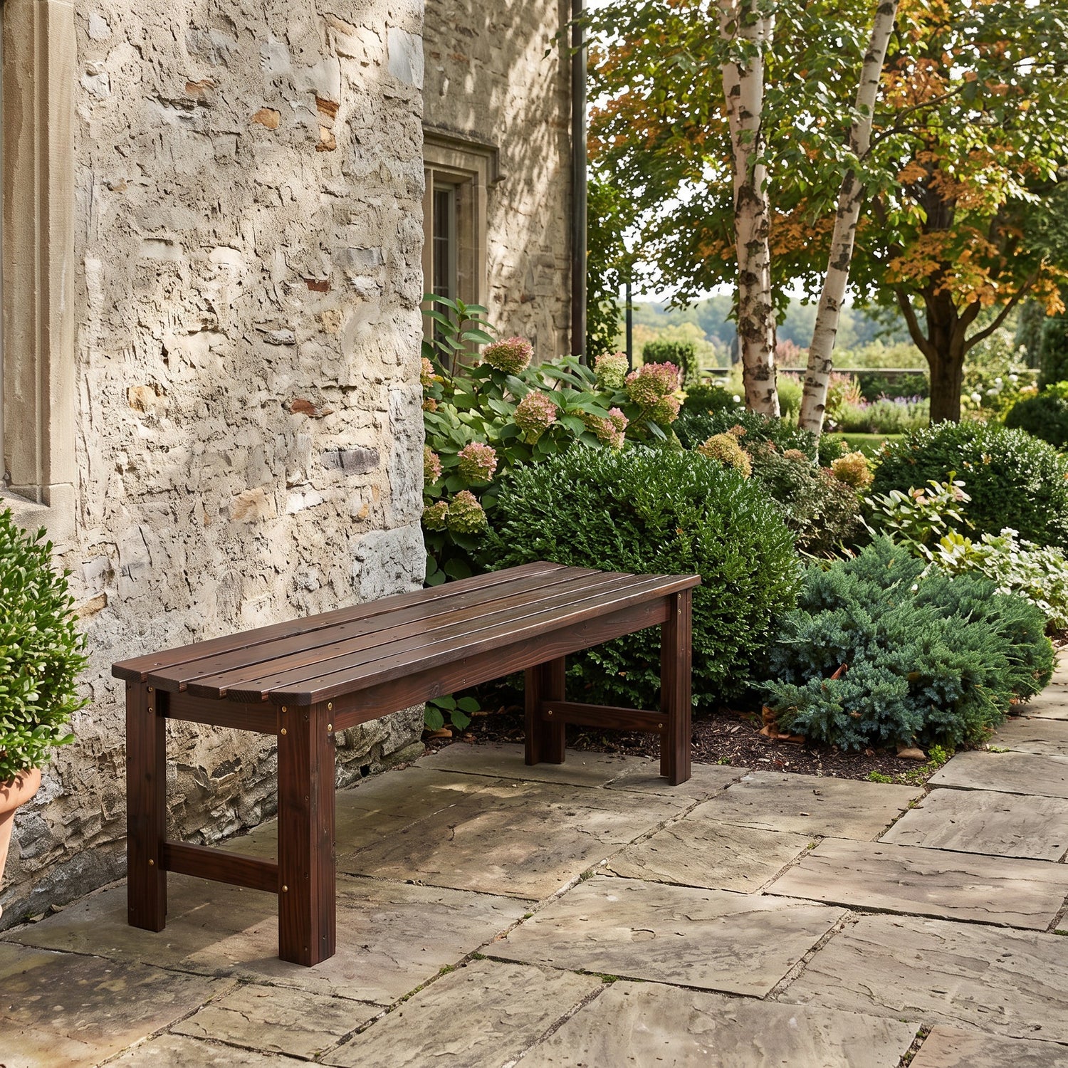 A dark brown wooden outdoor bench with a slatted seat and solid legs, positioned on a stone patio next to a rustic stone wall and a landscaped garden with green shrubs and trees. 