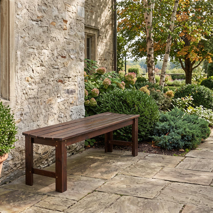 A dark brown wooden outdoor bench with a slatted seat and solid legs, positioned on a stone patio next to a rustic stone wall and a landscaped garden with green shrubs and trees. 