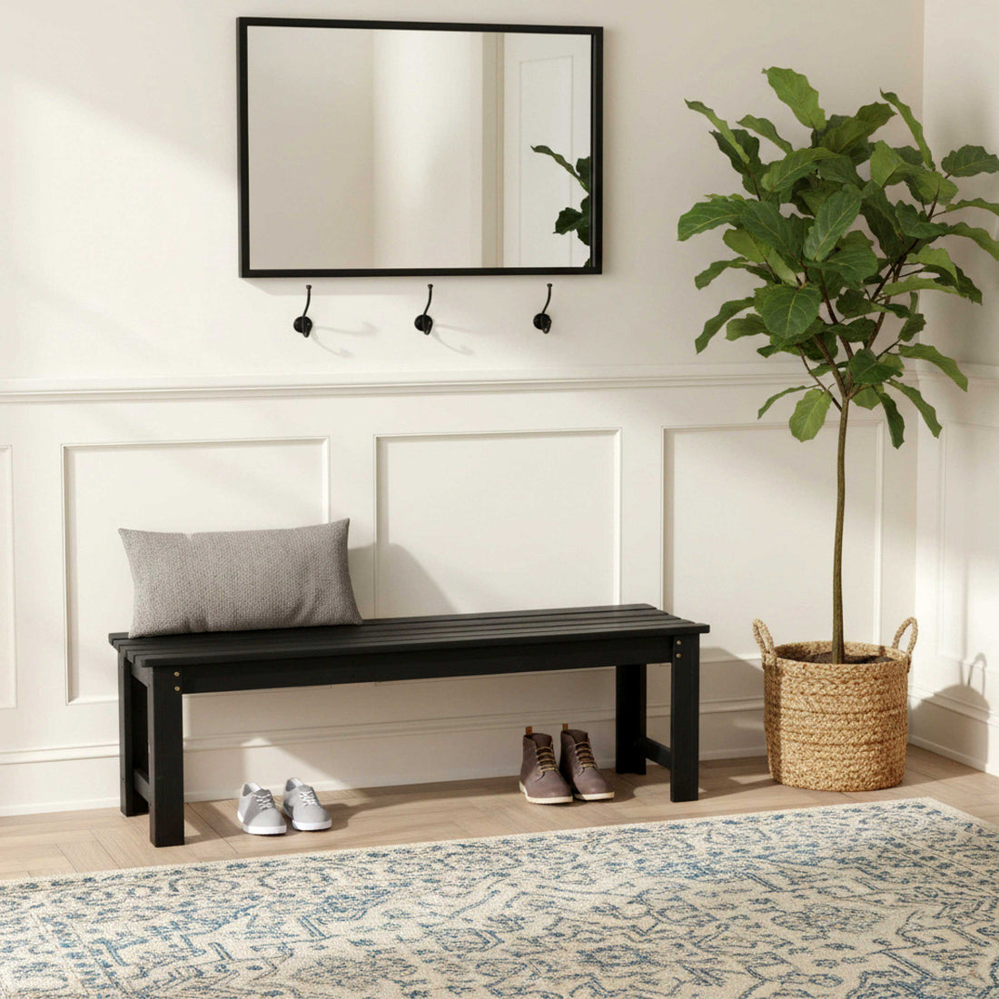 A black wooden entryway bench with a gray throw pillow, styled in a bright entryway with a white paneled wall, a black-framed mirror, wall hooks, a potted fiddle-leaf fig plant, shoes, and a patterned rug. 