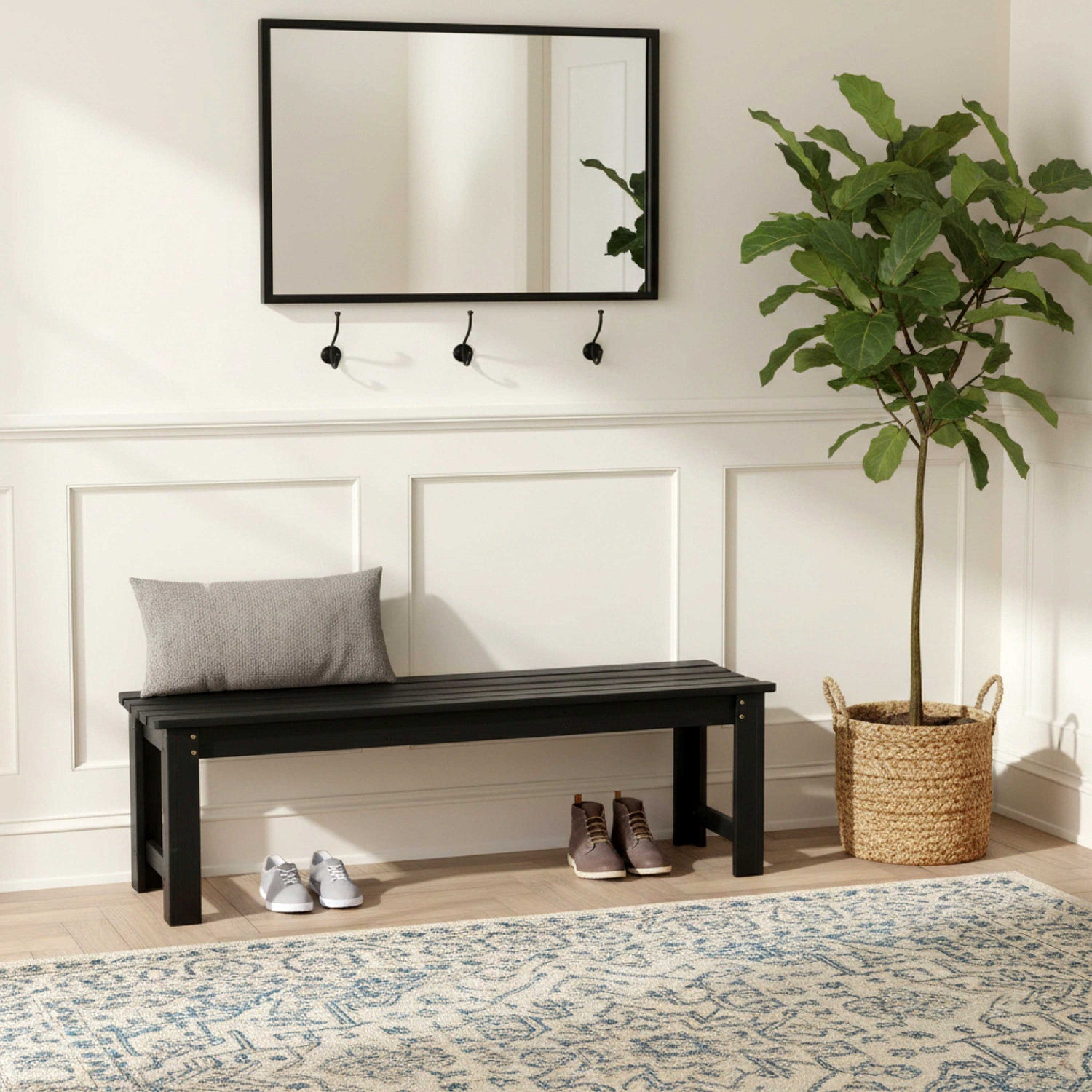 A black wooden entryway bench with a gray throw pillow, styled in a bright entryway with a white paneled wall, a black-framed mirror, wall hooks, a potted fiddle-leaf fig plant, shoes, and a patterned rug. 