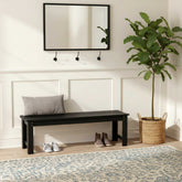 A black wooden entryway bench with a gray throw pillow, styled in a bright entryway with a white paneled wall, a black-framed mirror, wall hooks, a potted fiddle-leaf fig plant, shoes, and a patterned rug. 