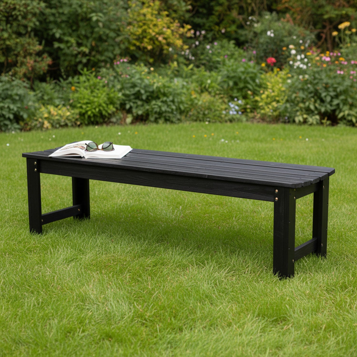 A long black outdoor bench with a slatted wooden top and sturdy legs, placed on a green lawn in a garden setting. An open book and sunglasses rest on the bench. 
