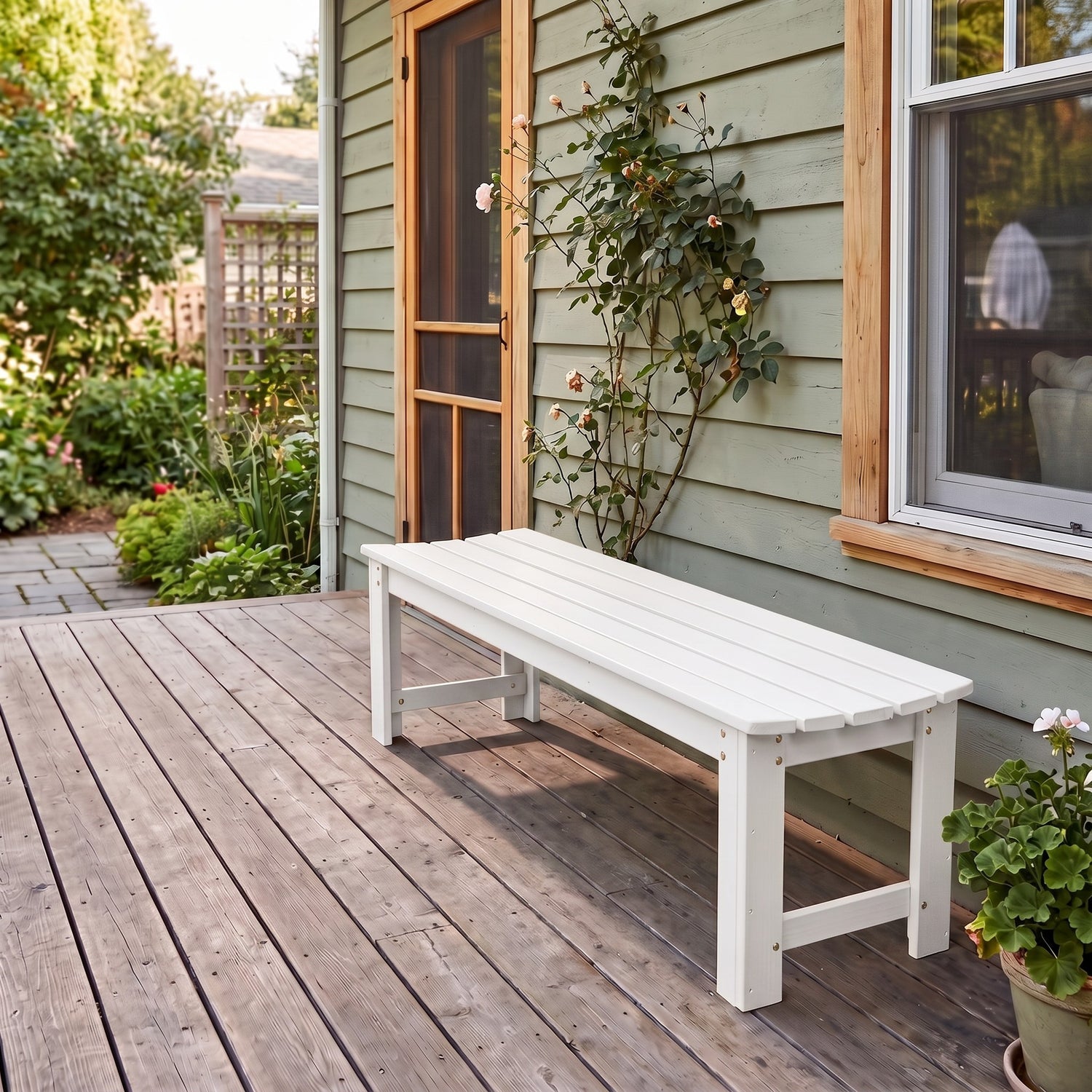 A white outdoor garden bench placed on a wooden deck beside a house with pale green siding, a window, and climbing roses.  