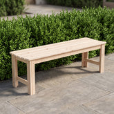 A light natural wood outdoor bench with a slatted seat and solid legs, positioned on a stone patio in front of a green hedge.  