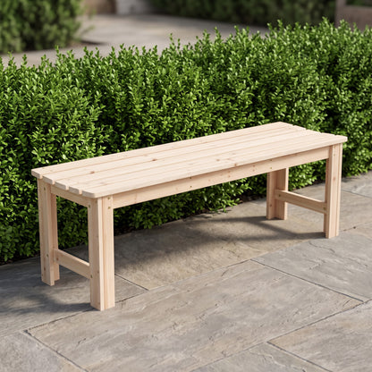 A light natural wood outdoor bench with a slatted seat and solid legs, positioned on a stone patio in front of a green hedge.  