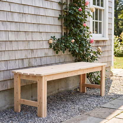 A light natural wood garden bench placed outdoors against a gray shingled house wall, with a climbing rose vine and pink flowers above it, on a gravel surface beside a stone walkway. 