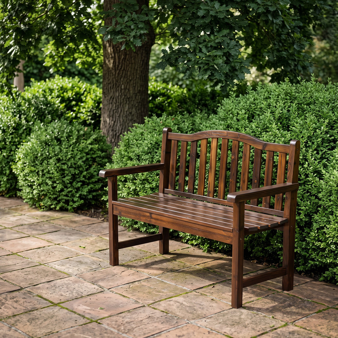 A dark brown wooden garden bench with slatted arms and backrest, placed on a stone patio in front of green shrubs and trees. 