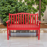 A bright red wooden garden bench with armrests and vertical slats, positioned on a light tiled patio in front of a stone wall and green ivy. 