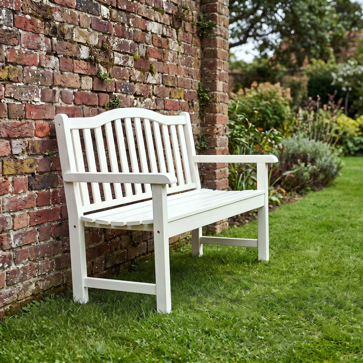 A white outdoor slatted bench with armrests, positioned on a green lawn next to a red brick wall and garden plants. 