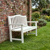 A white outdoor slatted bench with armrests, positioned on a green lawn next to a red brick wall and garden plants. 