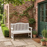 A white wooden garden bench with a slatted back and seat, placed on a stone patio in front of a red brick wall with climbing green vines and a dark green front door. 