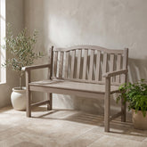 A single wooden garden bench with a slatted back and seat, shown in a softly lit patio setting with potted plants and a textured wall. 