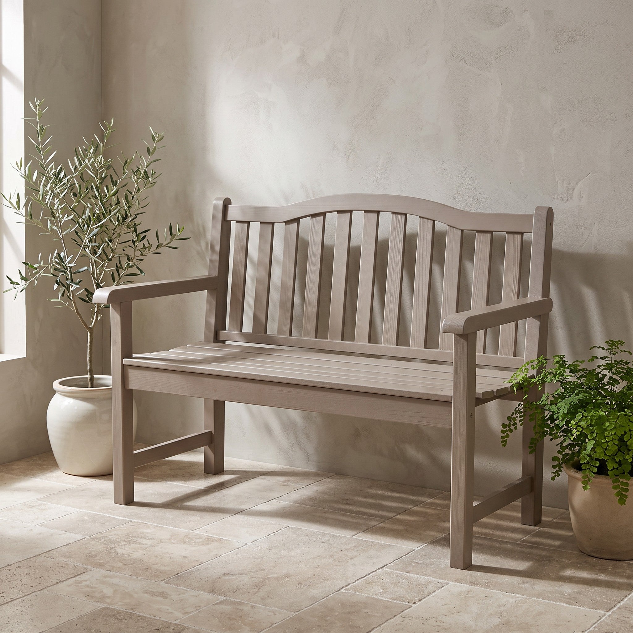 A single wooden garden bench with a slatted back and seat, shown in a softly lit patio setting with potted plants and a textured wall. 