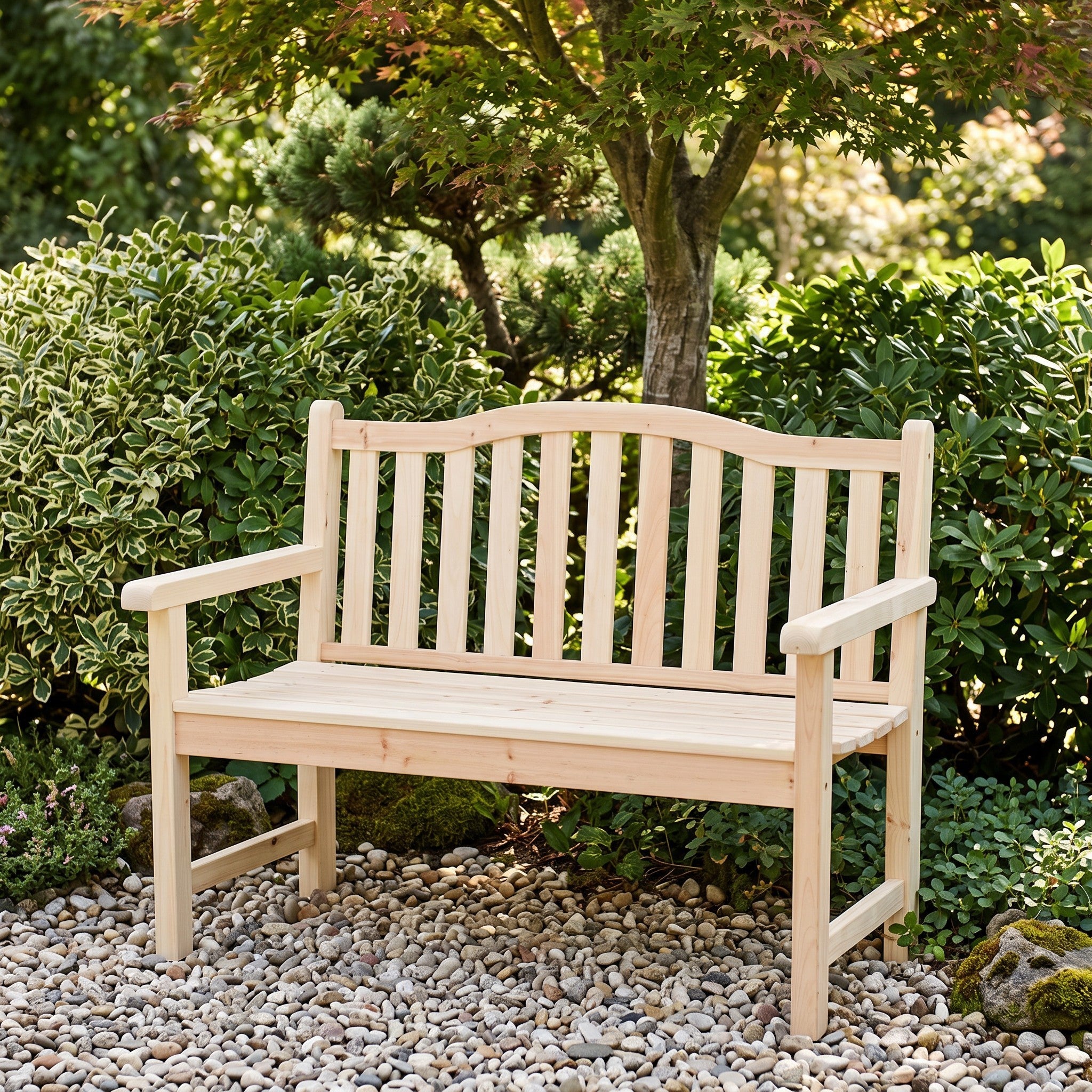 A light natural wood garden bench with a slatted backrest and armrests, placed on a bed of white gravel in a landscaped garden with shrubs and trees. 
