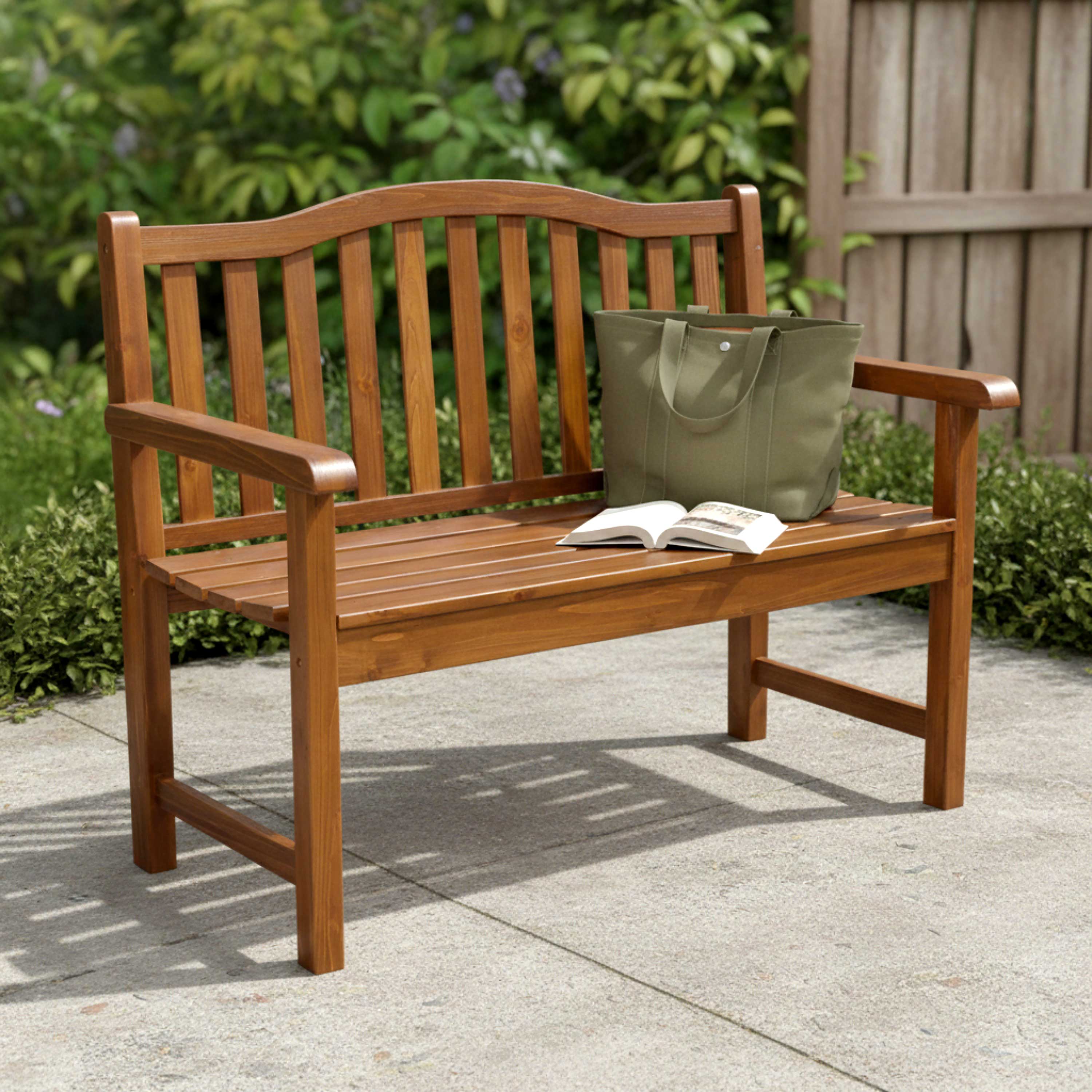 A wooden outdoor bench with a slatted back and seat, shown on a patio with a green tote bag and an open book placed on it. Lush green plants and a wooden fence are in the background. 