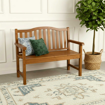 A wooden garden bench with a slatted back and seat, shown in a styled room setting with two decorative throw pillows, a patterned rug, and a potted tree. 