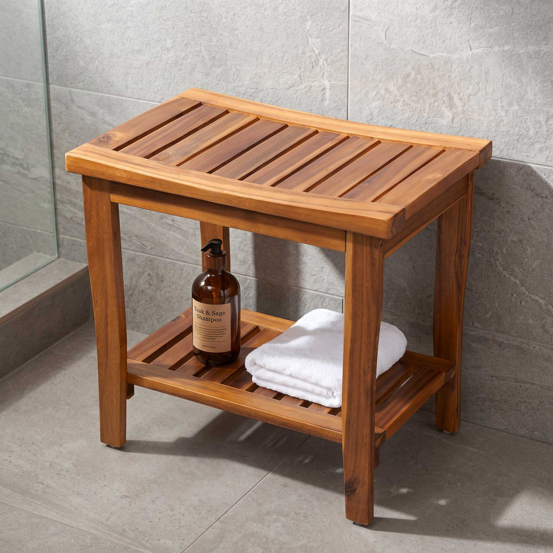 A wooden bathroom stool with a slatted top and lower shelf, shown in a tiled bathroom setting. A brown pump bottle and a folded white towel are placed on the shelf. 