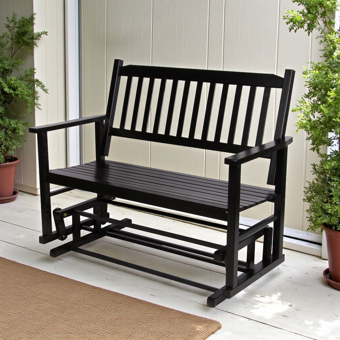 A black wooden outdoor porch bench with a slatted backrest and armrests, shown on a light porch with potted plants and a beige wall behind it. 