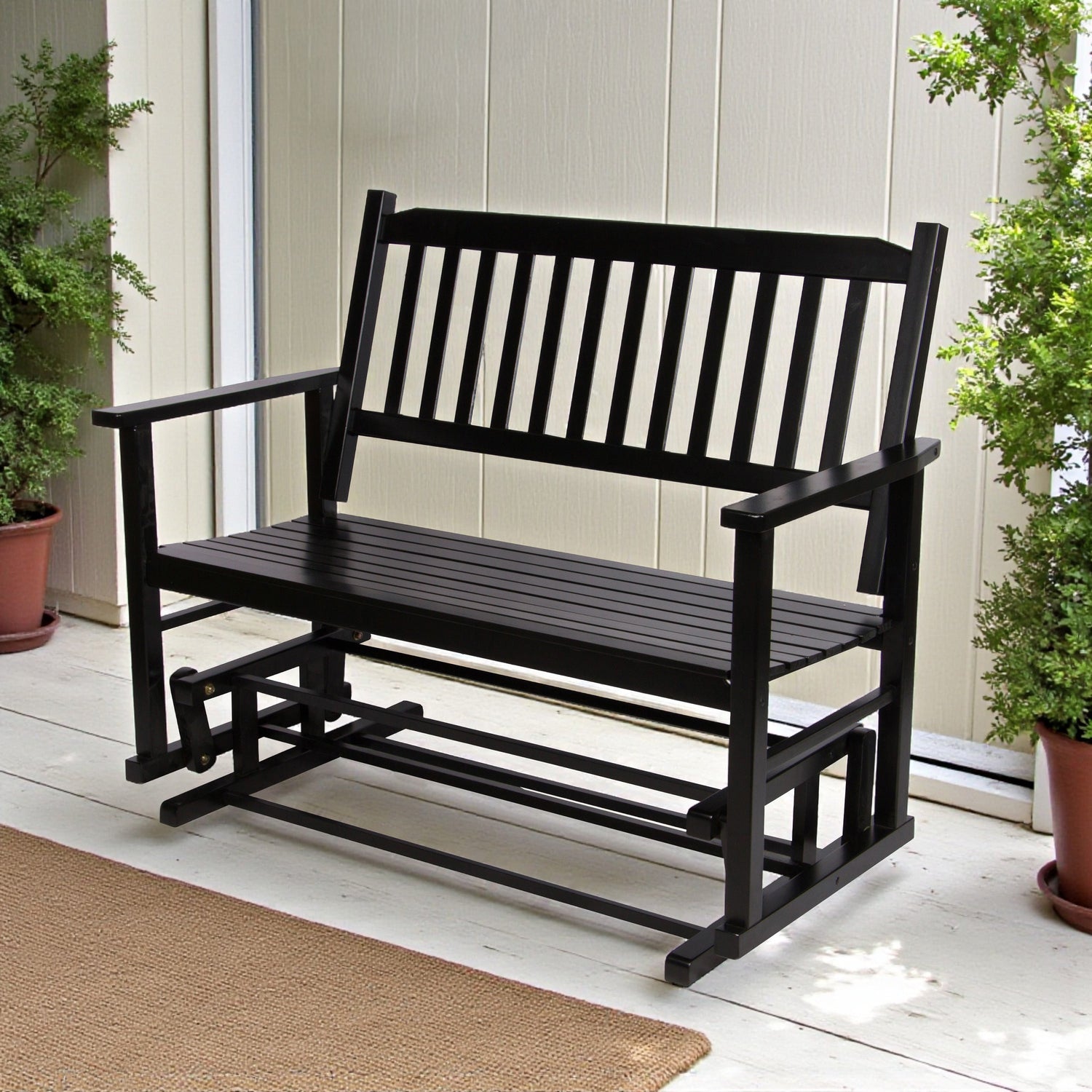 A black wooden outdoor porch bench with a slatted backrest and armrests, shown on a light porch with potted plants and a beige wall behind it. 
