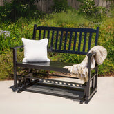 A black outdoor rocking bench with a white textured throw pillow and a beige knit blanket draped over the arm, set on a concrete patio with a green garden background. 
