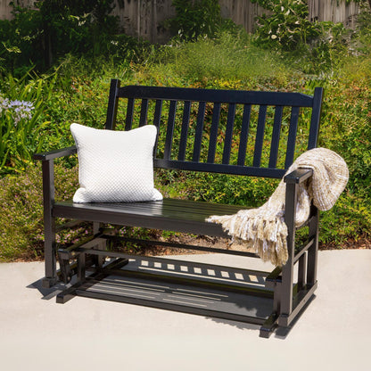 A black outdoor rocking bench with a white textured throw pillow and a beige knit blanket draped over the arm, set on a concrete patio with a green garden background. 