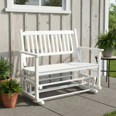 A white wooden porch rocking bench with armrests, shown outdoors on a patio in front of a light gray wood-paneled wall with potted plants nearby. 