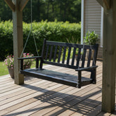 A black wooden porch swing hanging from chains on a wooden deck, with a garden and house siding in the background. 