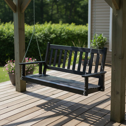 A black wooden porch swing hanging from chains on a wooden deck, with a garden and house siding in the background. 