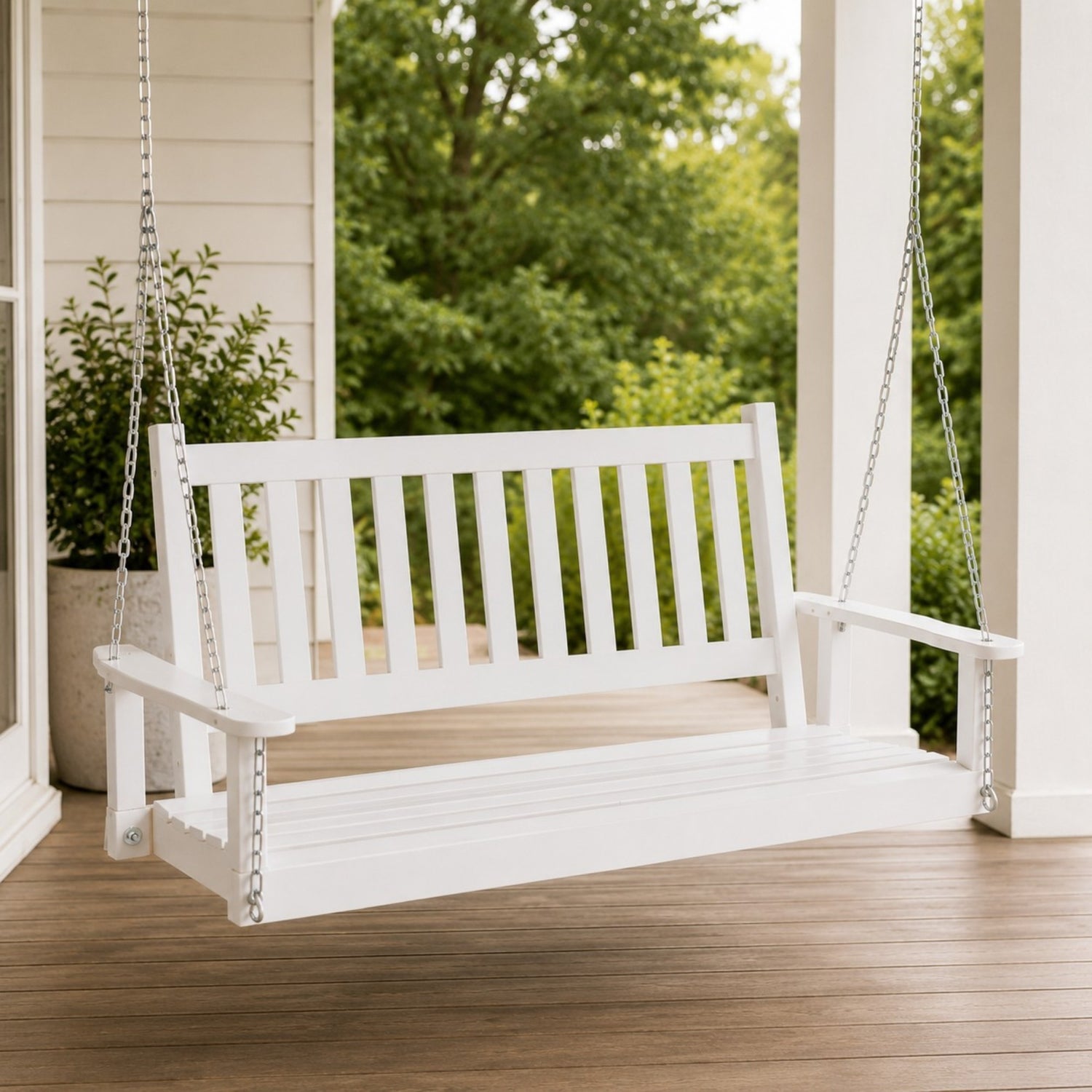 A white wooden porch swing hanging from silver chains on a covered porch with a wooden floor and green trees in the background. 