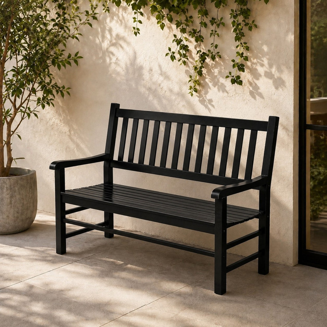 A black outdoor bench with a slatted back and seat, placed on a patio against a stucco wall with climbing greenery and a potted plant nearby. 