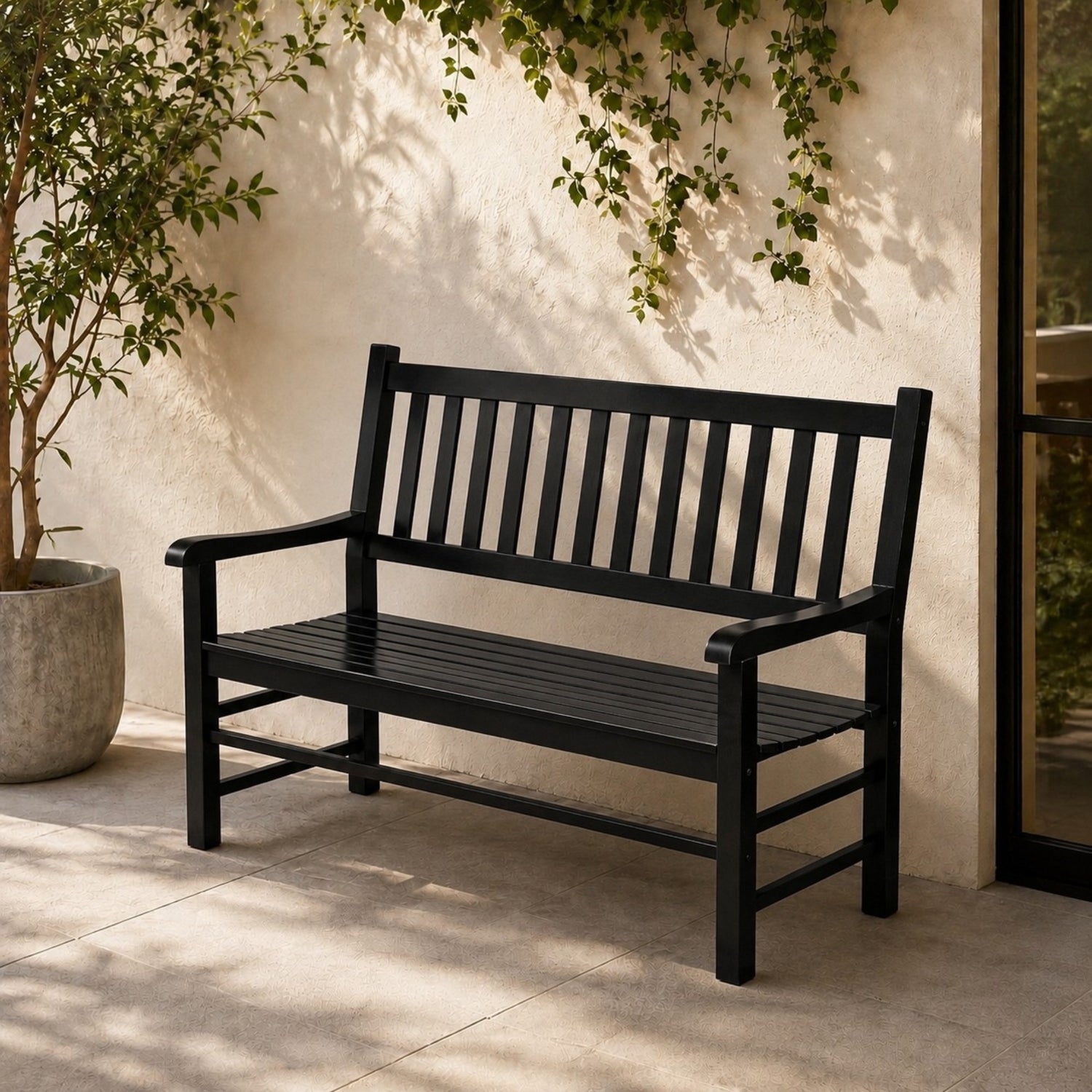 A black outdoor bench with a slatted back and seat, placed on a patio against a stucco wall with climbing greenery and a potted plant nearby. 