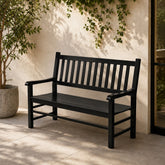 A black outdoor bench with a slatted back and seat, placed on a patio against a stucco wall with climbing greenery and a potted plant nearby. 