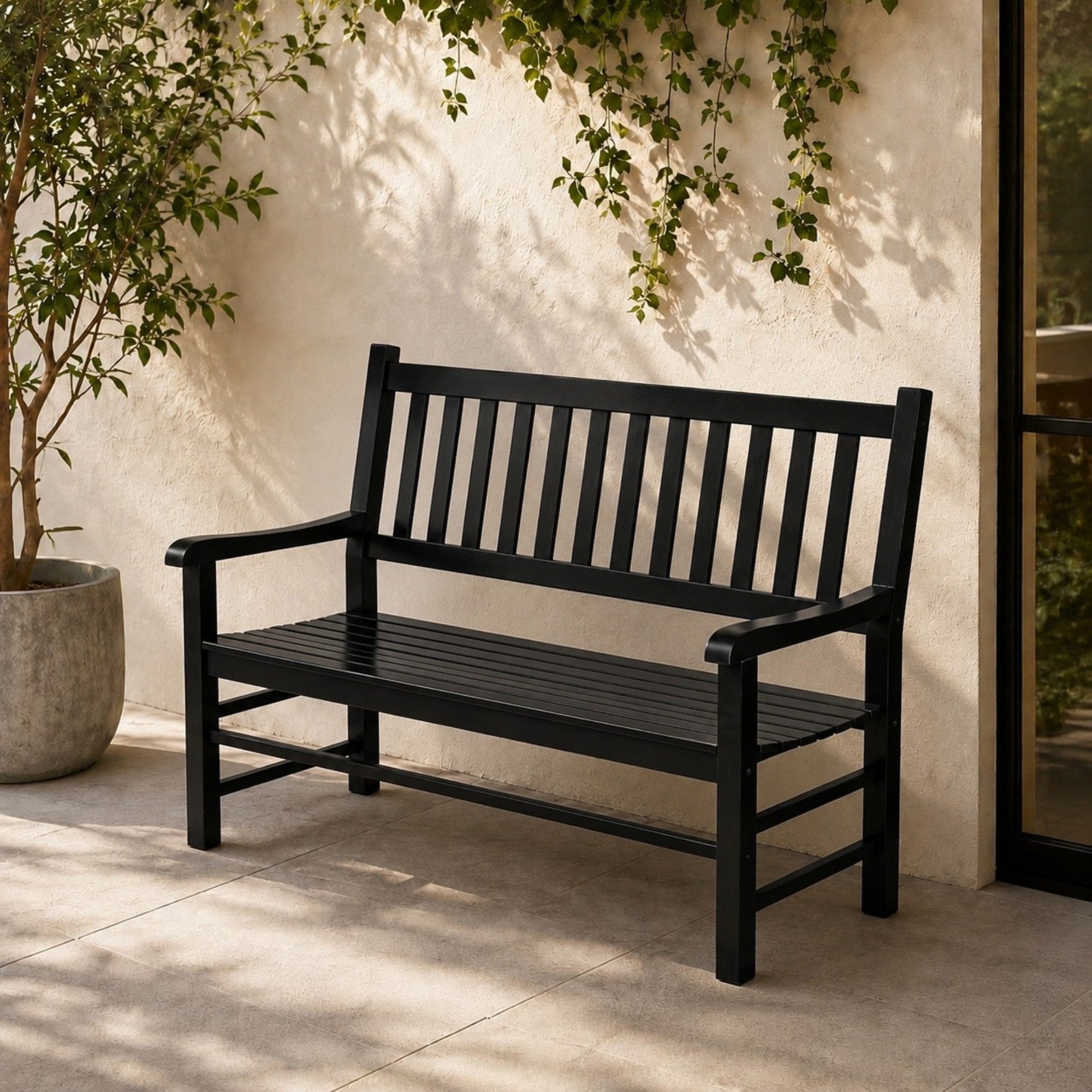 A black outdoor bench with a slatted back and seat, placed on a patio against a stucco wall with climbing greenery and a potted plant nearby. 