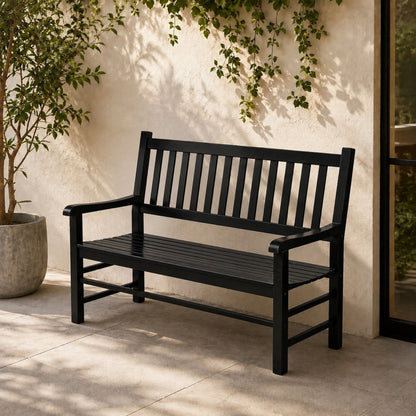 A black outdoor bench with a slatted back and seat, placed on a patio against a stucco wall with climbing greenery and a potted plant nearby. 