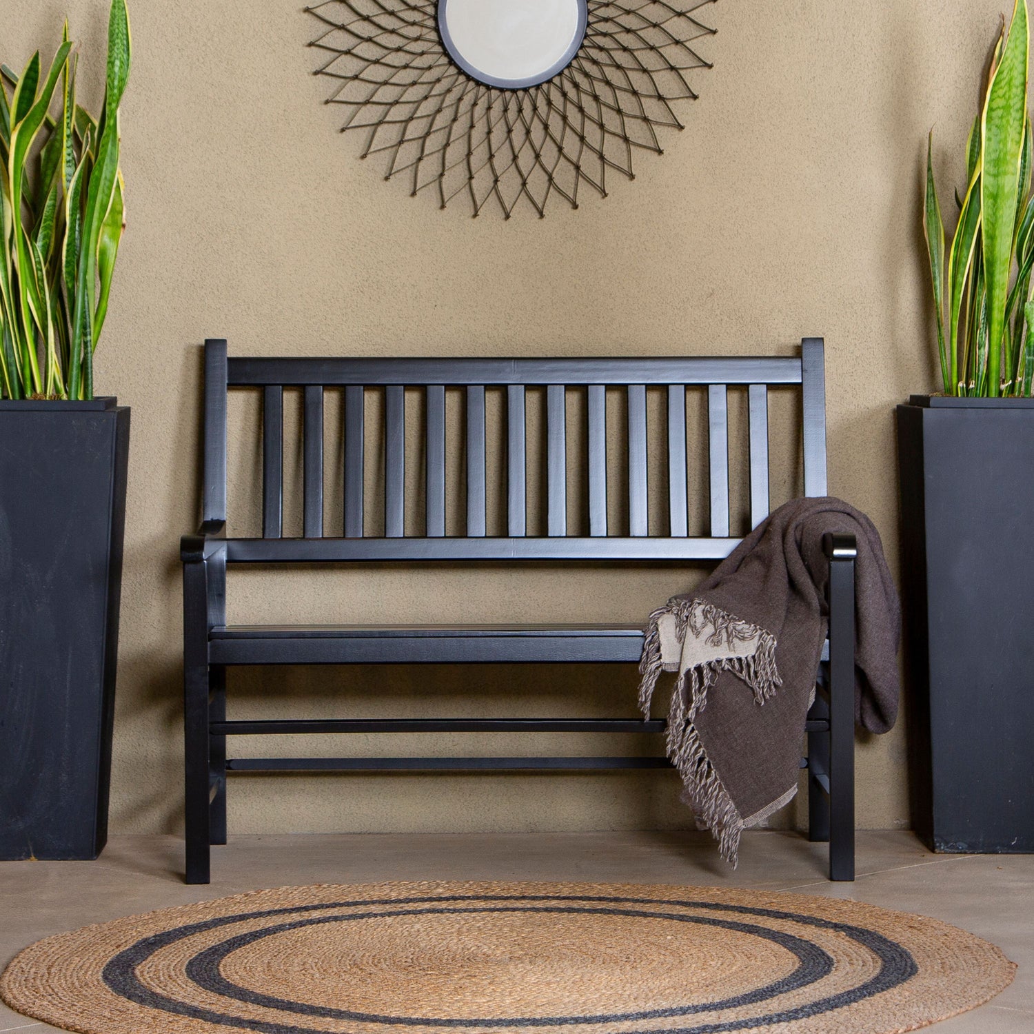 A black wooden slat bench is centered against a beige wall, with a brown throw blanket draped over one arm. Two tall black planters with green snake plants flank the bench, and a round woven rug sits in front. 