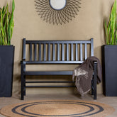 A black wooden slat bench is centered against a beige wall, with a brown throw blanket draped over one arm. Two tall black planters with green snake plants flank the bench, and a round woven rug sits in front. 